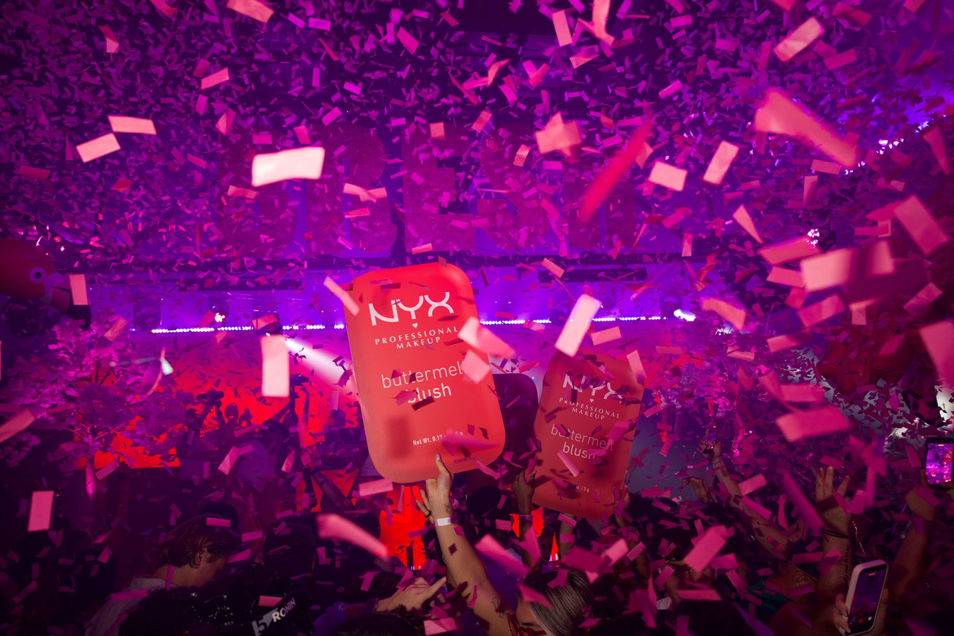 A cardboard cutout of NYX Buttermelt Blush product held above a crowd with pink light and confetti falling all around.