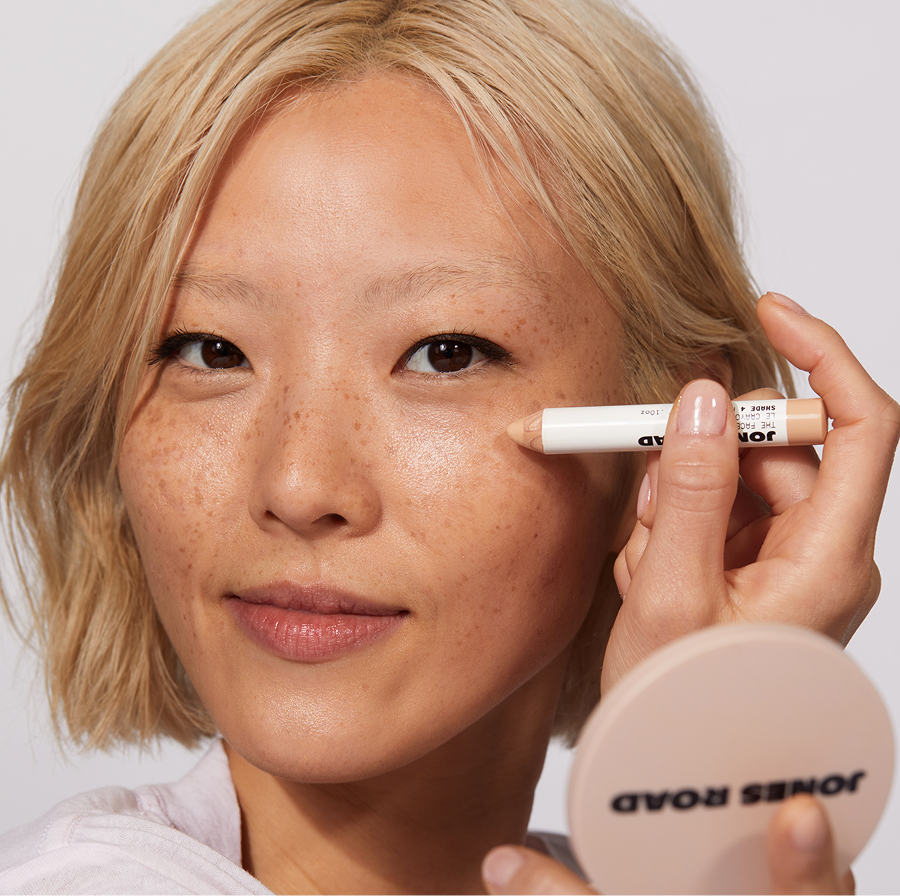 Model applying The Face Pencil under her eye while holding a Jones Road mirror