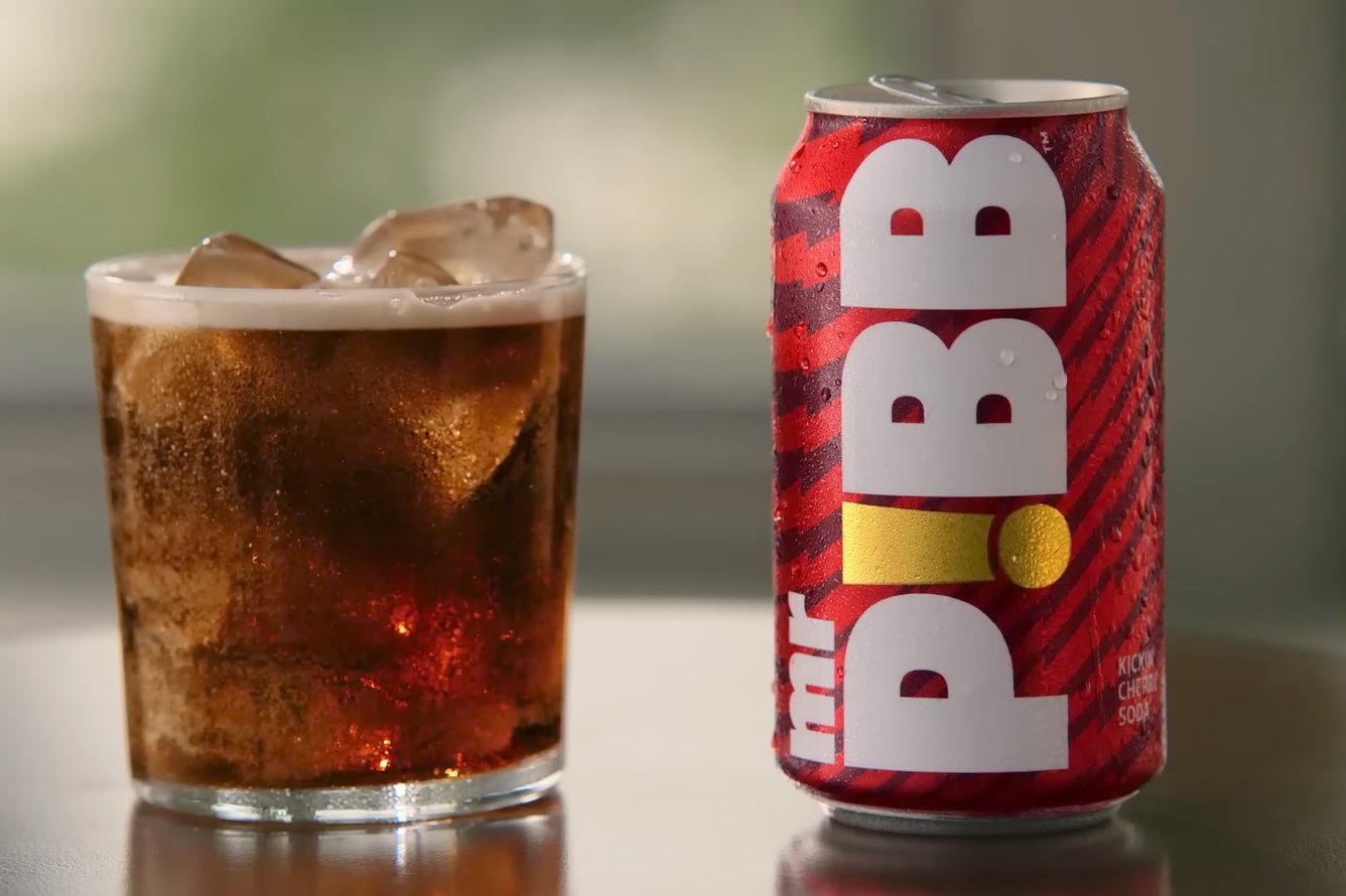 A transparent glass containing a brownish-red carbonated soda and ice cubes on the left, and a red Mr. Pibb can on the right