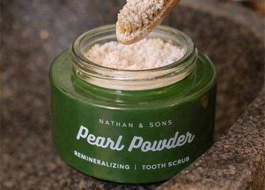 A close-up, slightly overhead view shows an open dark green glass jar filled with pale powder on a textured brown stone surface, with a wooden spoon l...