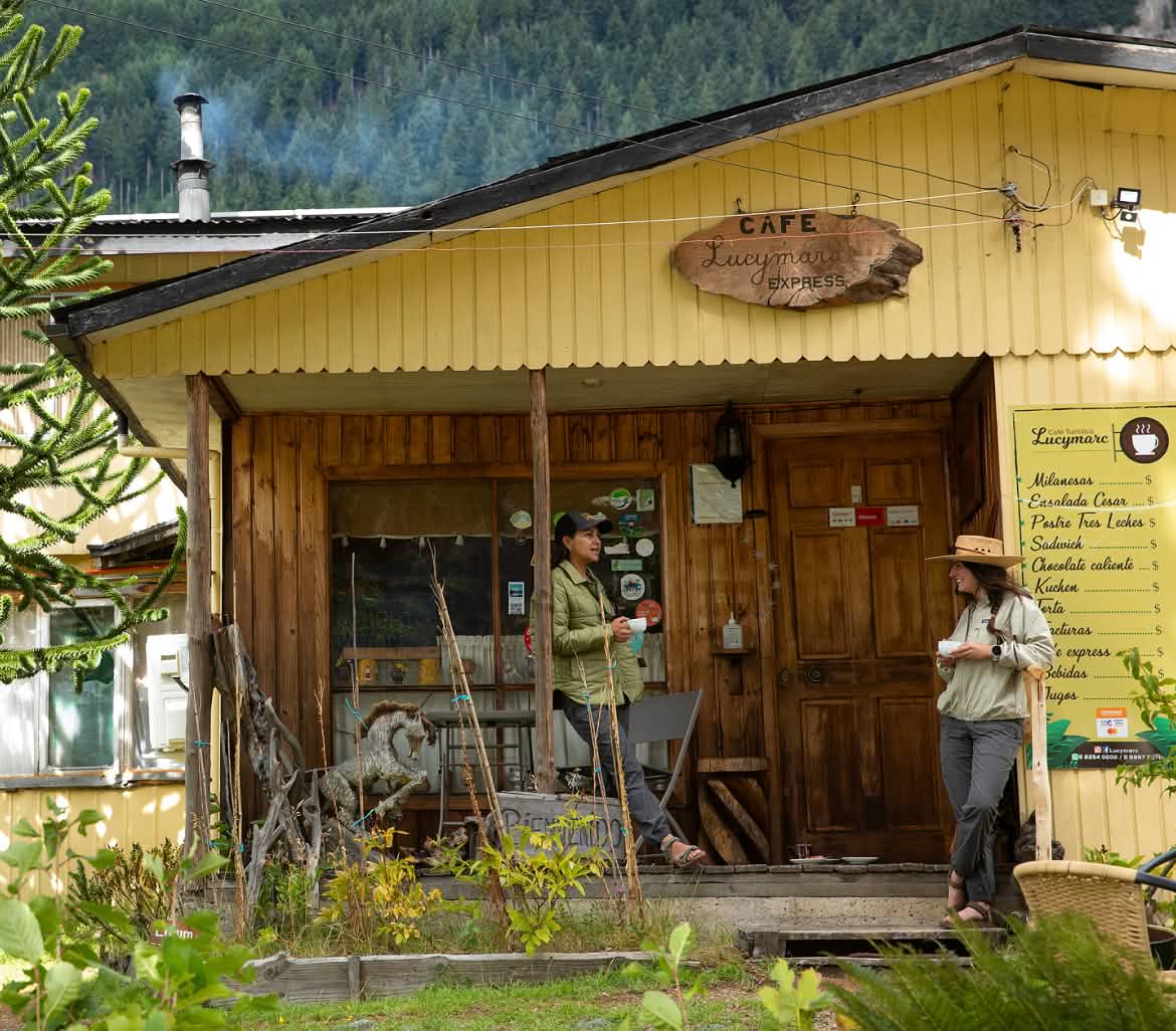 wo people stand outside a mountain cafe chatting and holding coffee cups.