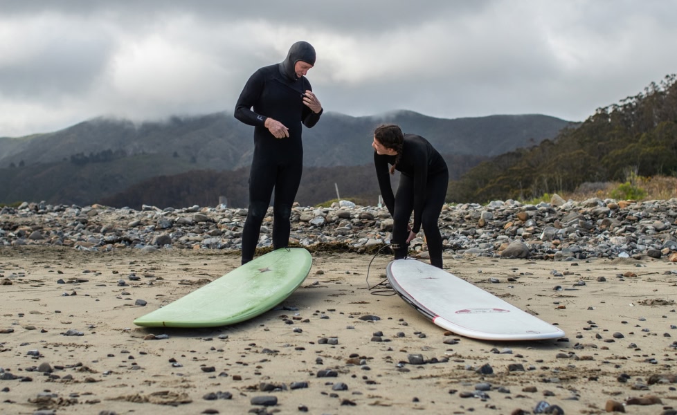 Two people wearing wetsuits stand on a beach with their longboards lying on the sand in front of them.