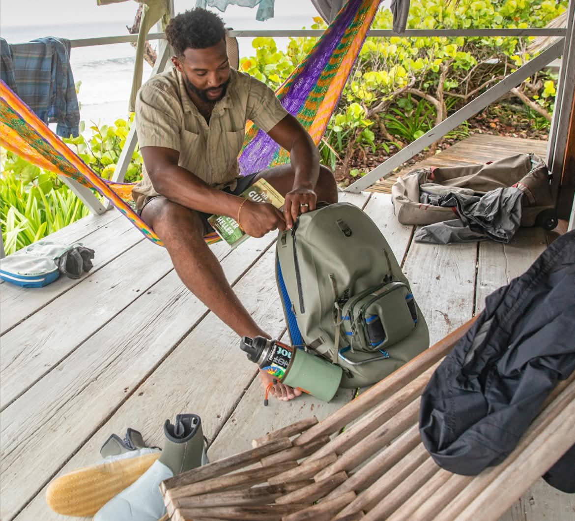 A person organizes gear in a backpack while sitting in a hammock on a porch overlooking a beach.
