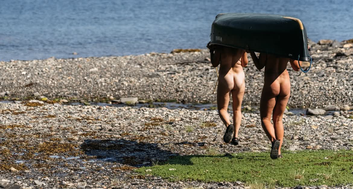 Patagonia. Two people who are not wearing clothes carry a canoe over their heads while walking toward a river.
