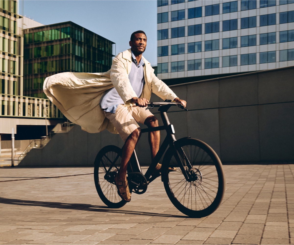 A man riding a black Cowboy bike in an urban setting, wearing a flowing light-coloured coat. The background features modern architecture with glass and concrete buildings.