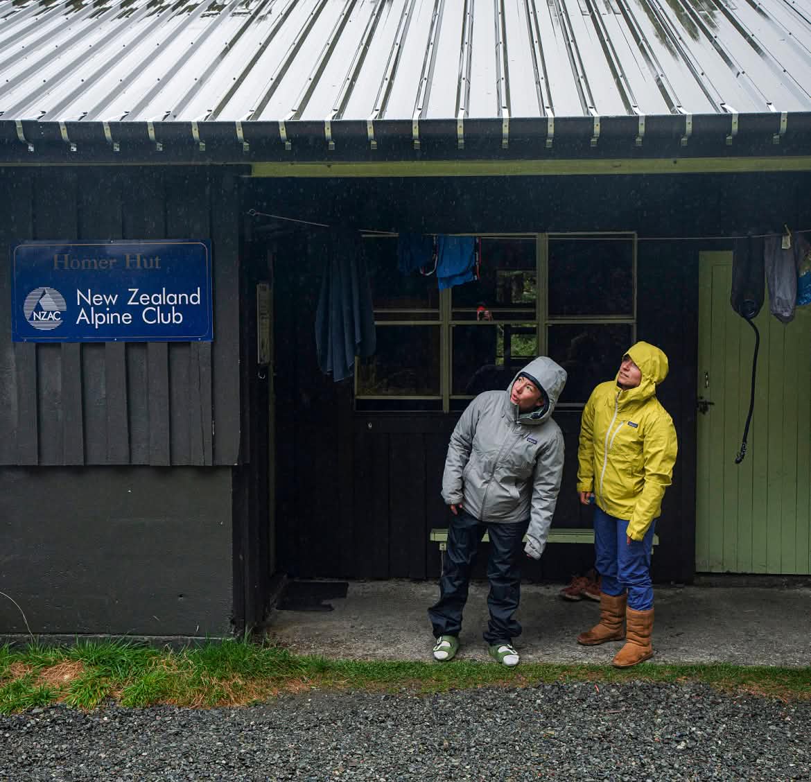 Two people wearing rain jackets peek up at the sky from under a porch roof overhang.