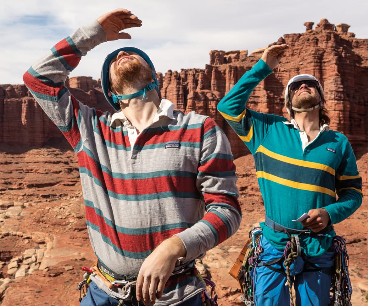 Two climbers wearing rugby shirts and climbing harnesses use their hands to block the sun while looking up at a wall route in Canyonlands National Park, Utah.