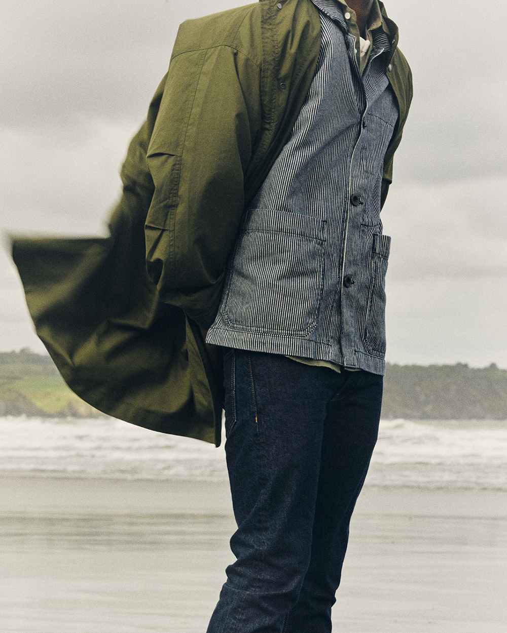 Image of woman on beach with sand covered hands. She wears Rinse Wash denim jeans, green shirt, and green sleeveless jumper.