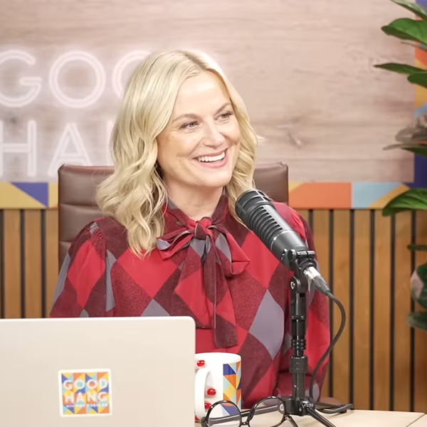 Amy Poehler smiles behind laptop and microphone, holding mug.