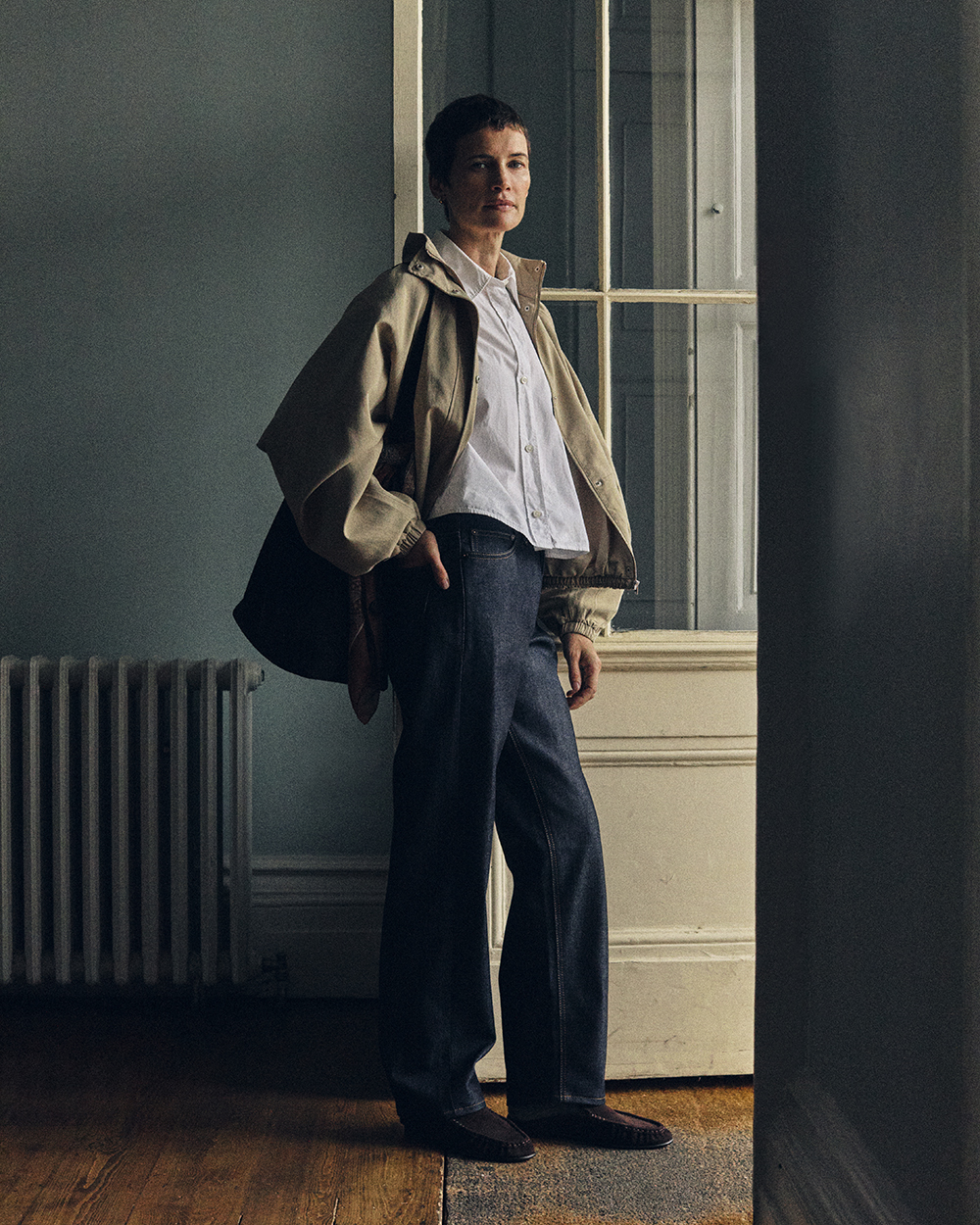 Man wearing dark selvedge jeans and a brown jacket beside a stone doorway.