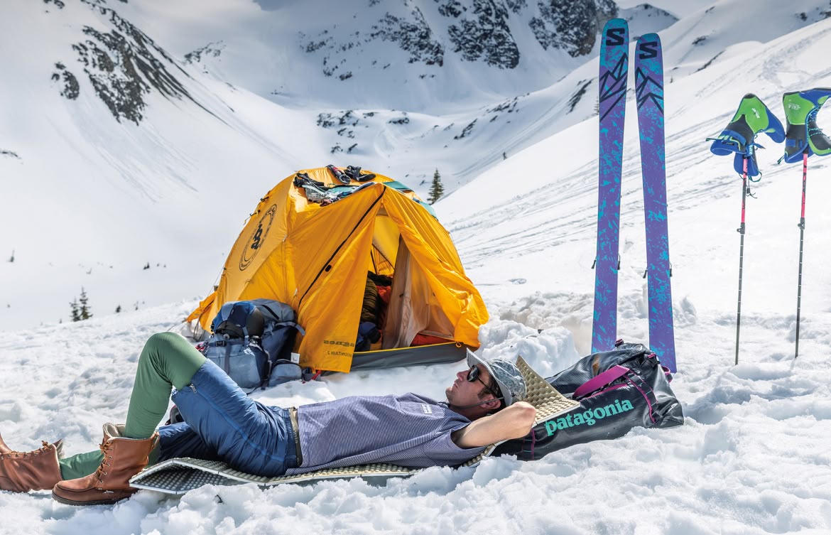 A backcountry skier lounging in the sun outside of their tent.