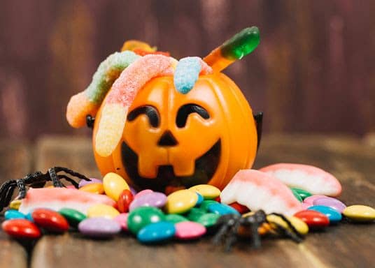 Close-up view of an orange pumpkin-shaped candy with black facial features and colorful gummy worms draped over it, surrounded by multicolored candy p...