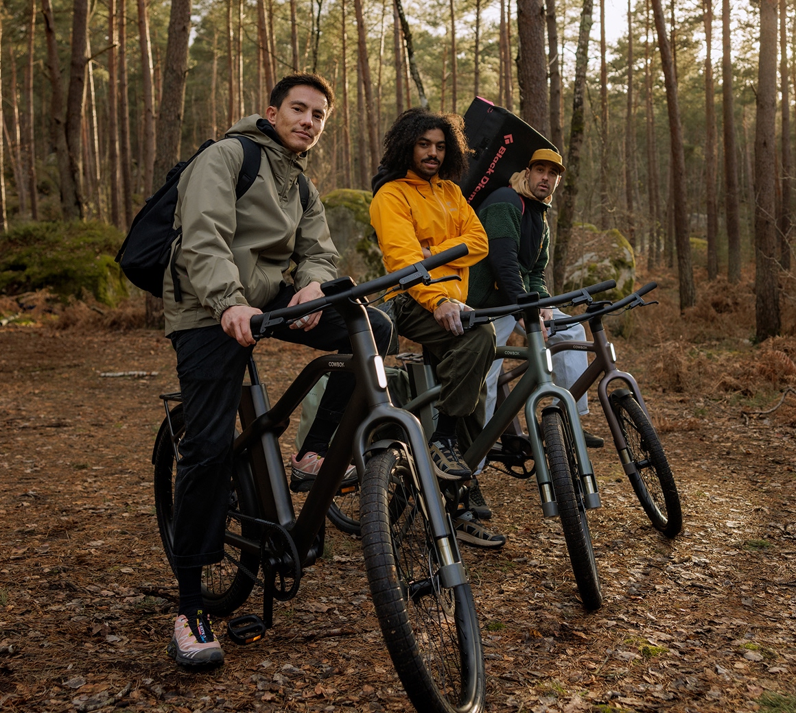 Three riders pause with their Cowboy Cross e-bikes on a pine forest trail covered in fallen leaves.