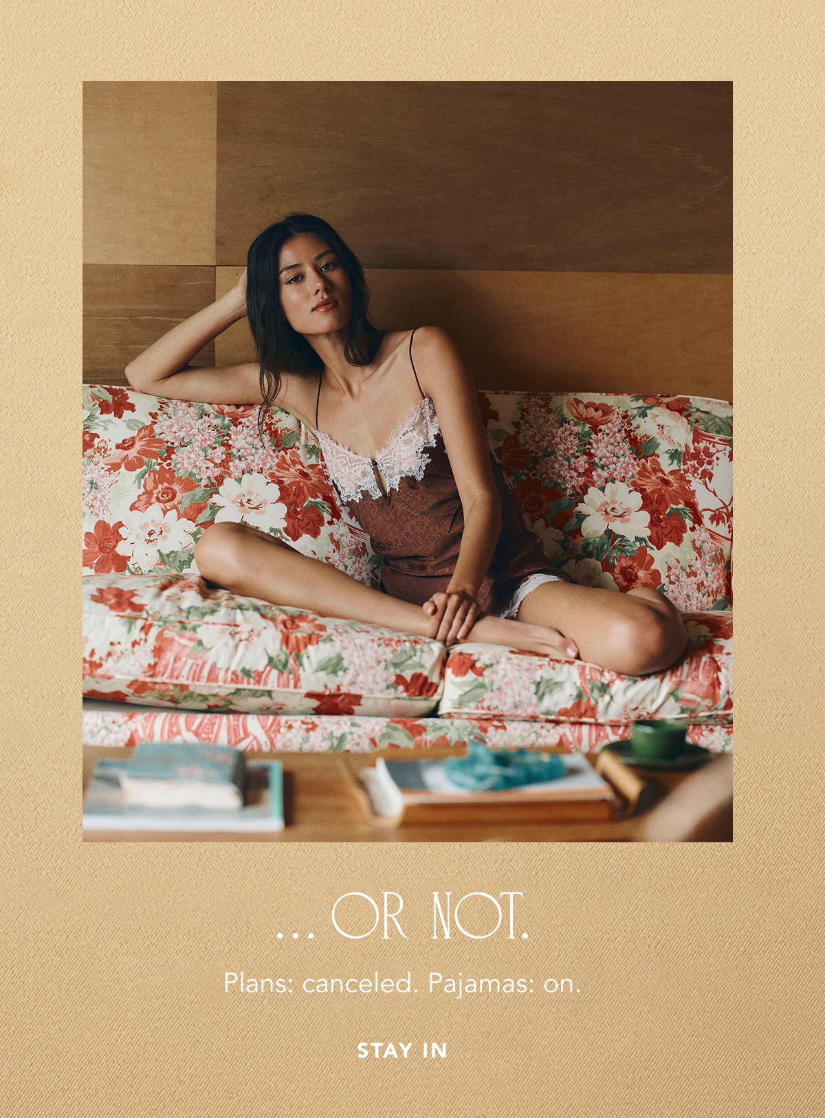 Model lounging on a red floral sofa wearing a brown satin slip dress trimmed with white lace, set against a warm beige background with text reading "... OR NOT. Plans: canceled. Pajamas: on. STAY IN".