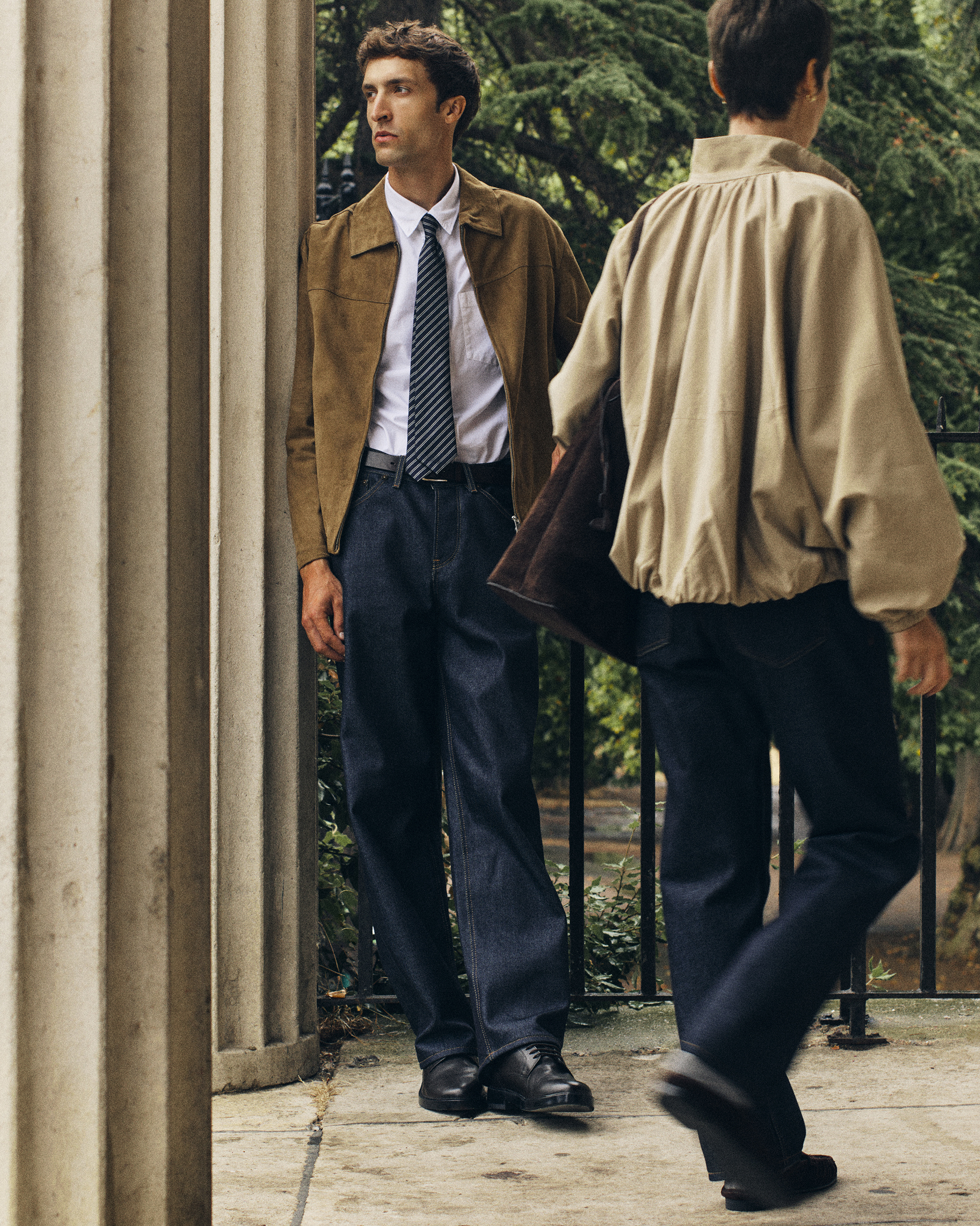Man leans against pillar wearing Super Zero Hiut jeans, black leather shoes, white shirt, tie, and brown suede jacket. Woman walks past wearing Super Zero Hiut jeans, cream jacket and brown leather bag.