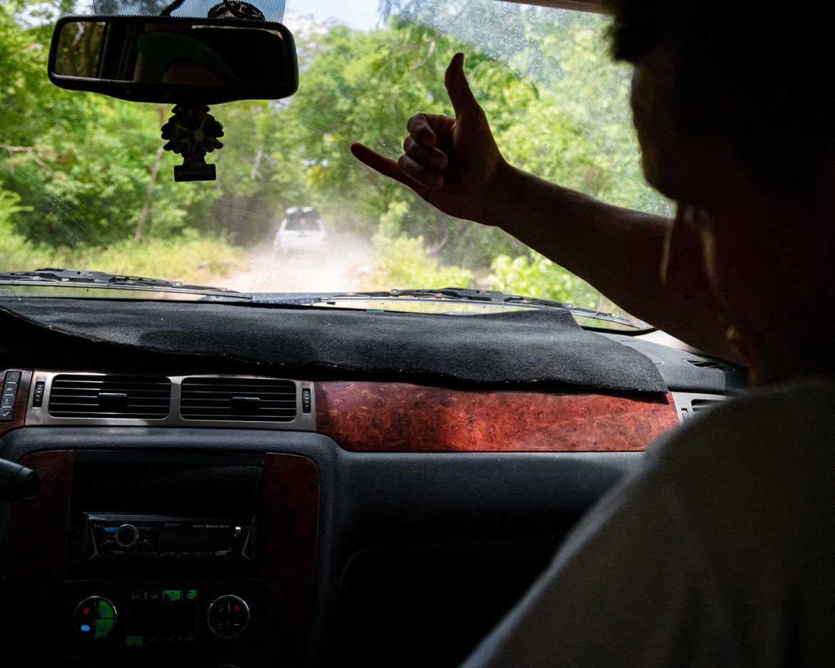 A photo taken from the backseat of a car following another car down a  dirt road. We see the driver&rsquo;s hand on the wheel and the left side of the passenger&rsquo;s  face. Passenger is making a &ldquo;shaka&rdquo; hand sign. 