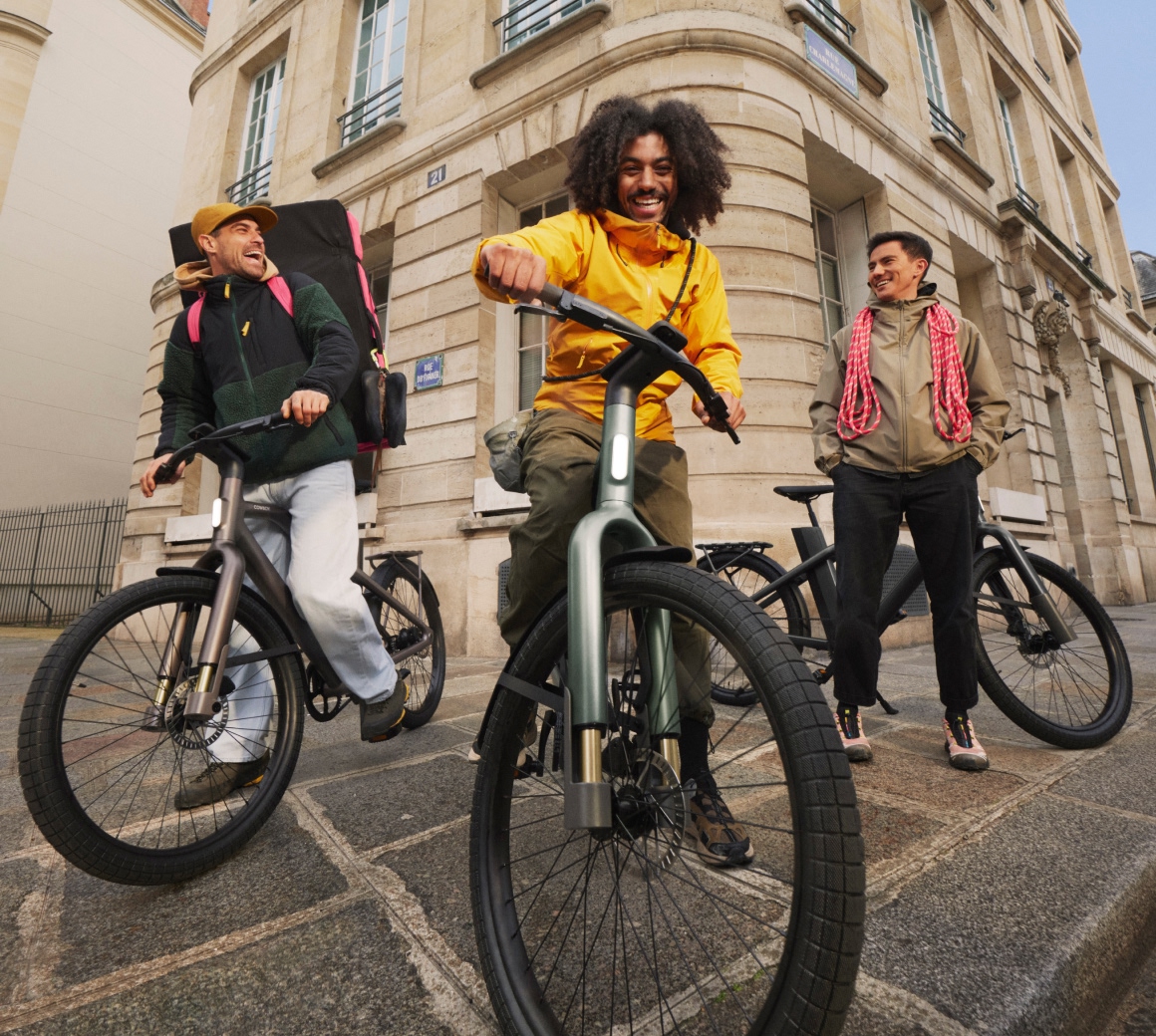 Three friends with Cowboy Cross e-bikes on a city street corner, one carrying a climbing rope, another a crash pad.