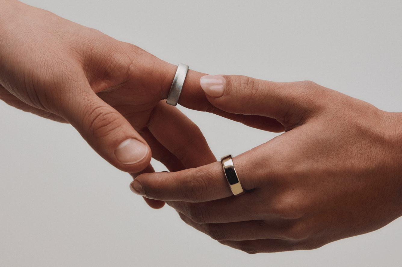 Two hands reach toward each other, each wearing Oura Rings, one silver and one gold