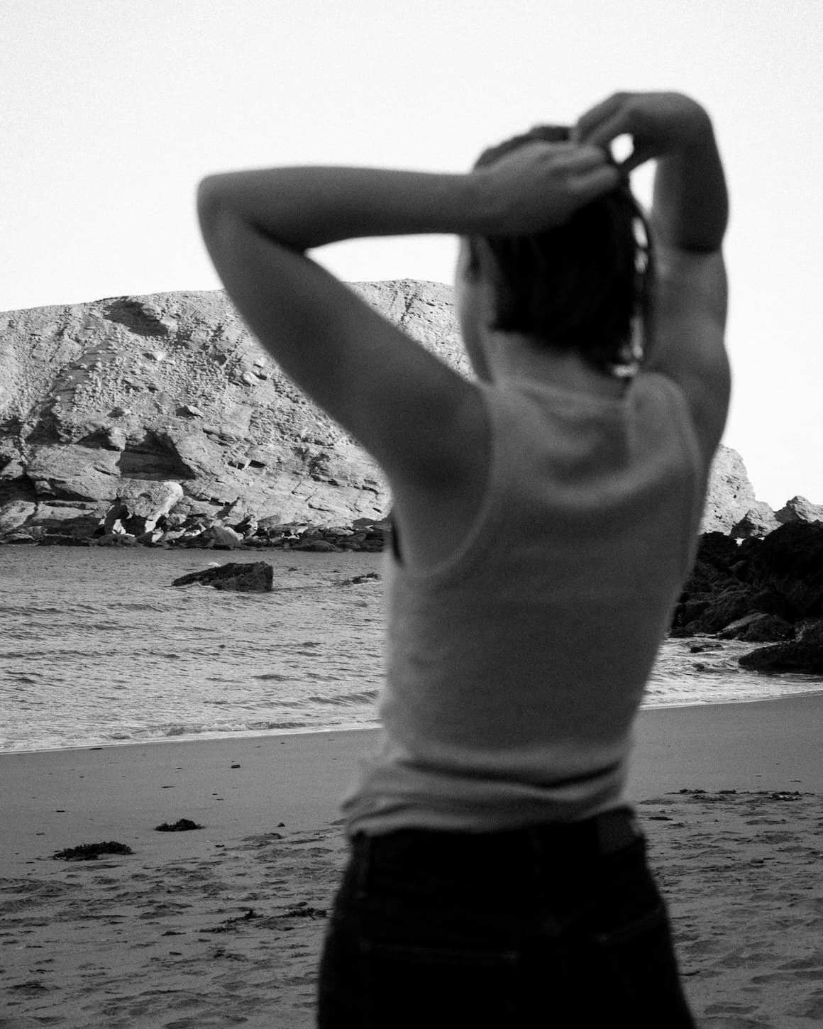 Woman stand on beach looking out to sea, with small rocky island in the background. She is slightly blurred in a white vest and jeans, with only waist up visible in the foreground.