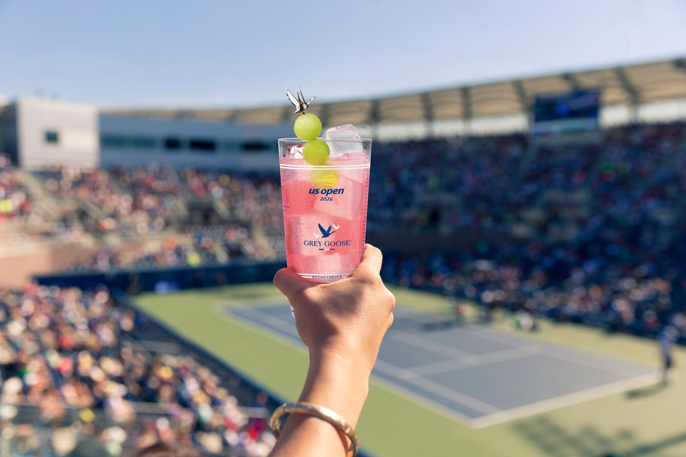 Hand holding a Grey Goose Honey Deuce in commemorative cup overlooking the courts at the 2026 US Open.