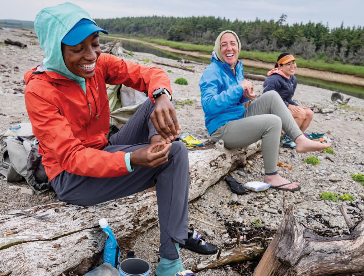 Three people wearing rain and running gear sit on a log on the pebbly bank of a river.