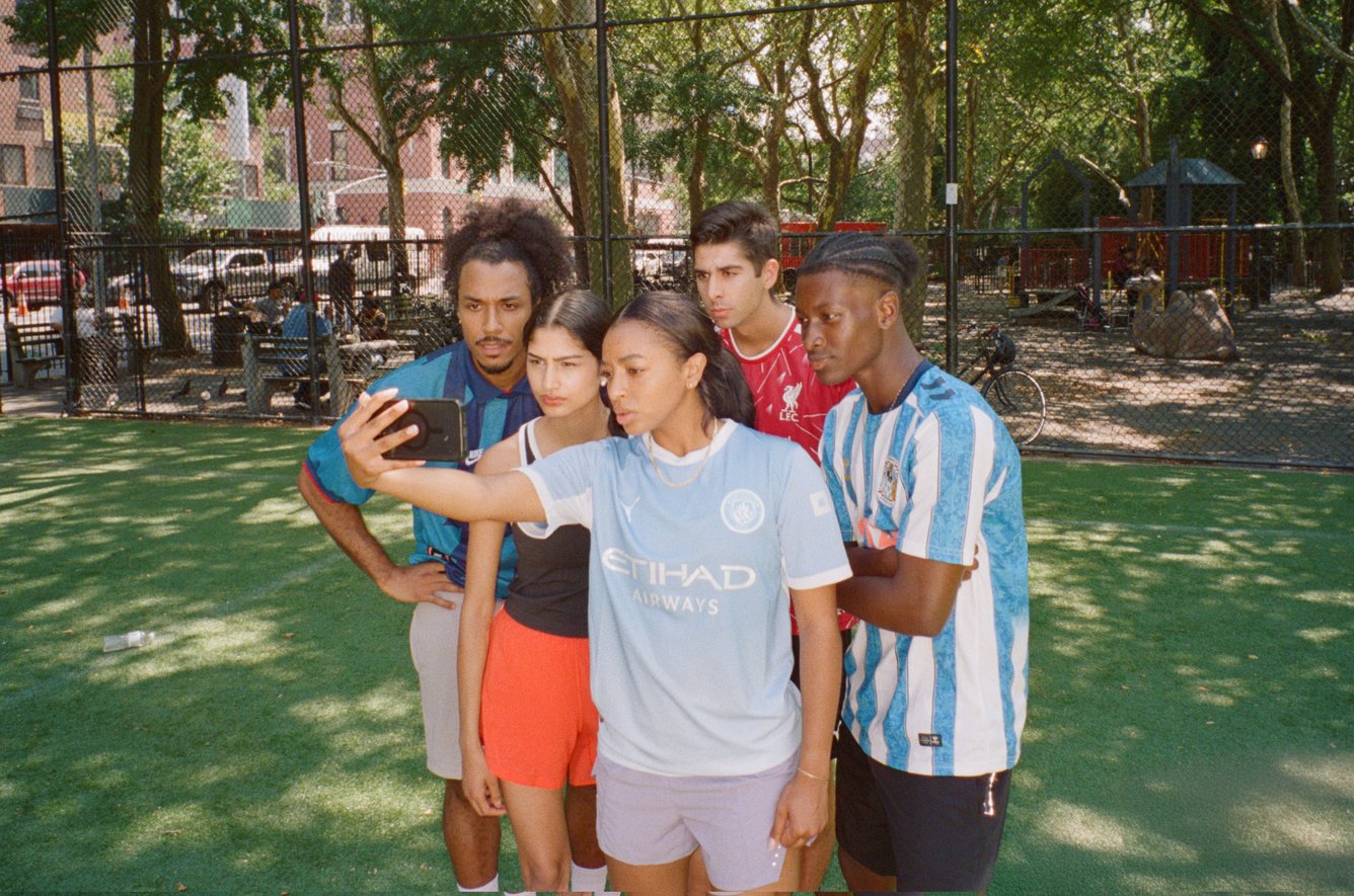 Fans in Premier League soccer jerseys watch a phone screen at a park.