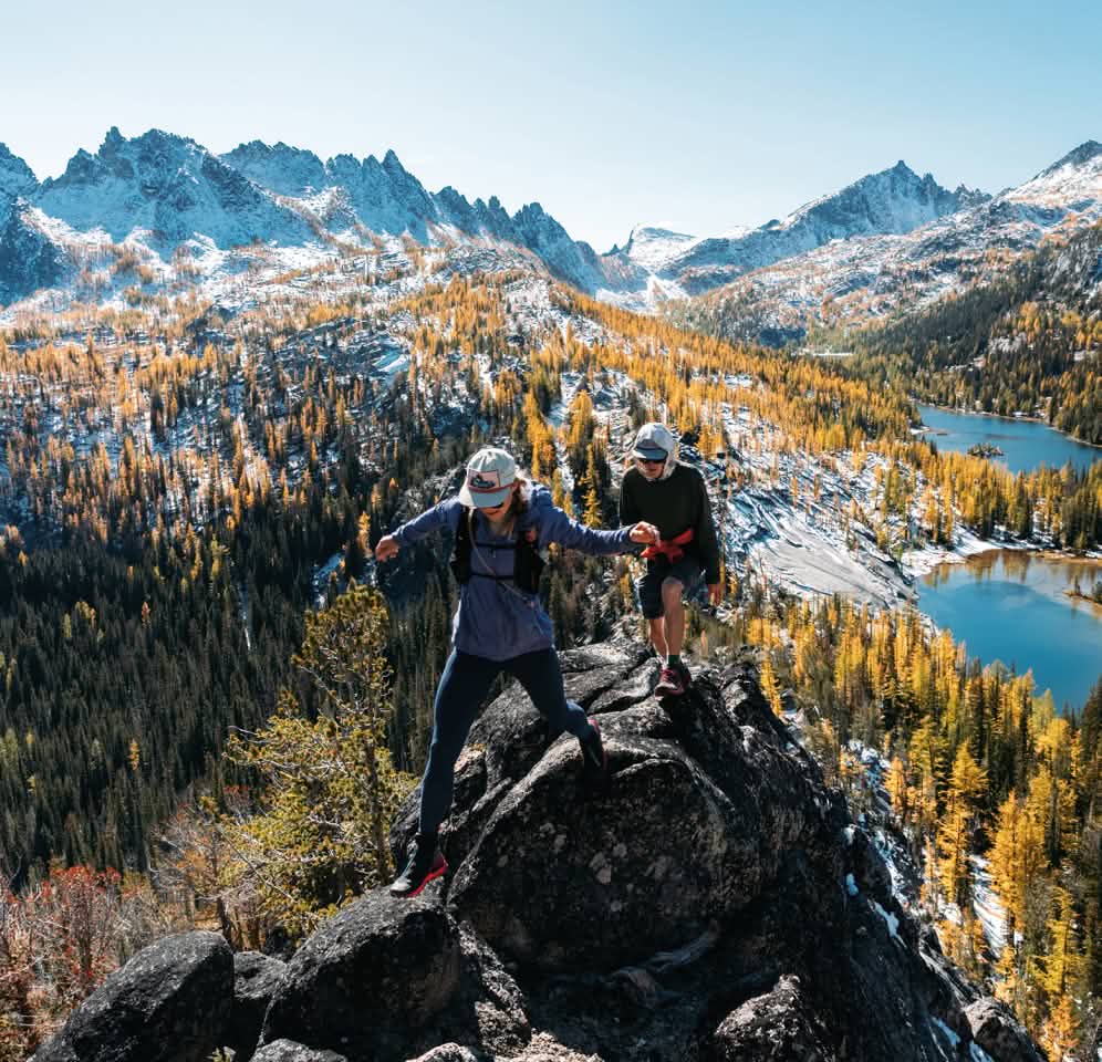 Two people on a rocky outcrop overlooking an alpine basin aglow with golden larch.