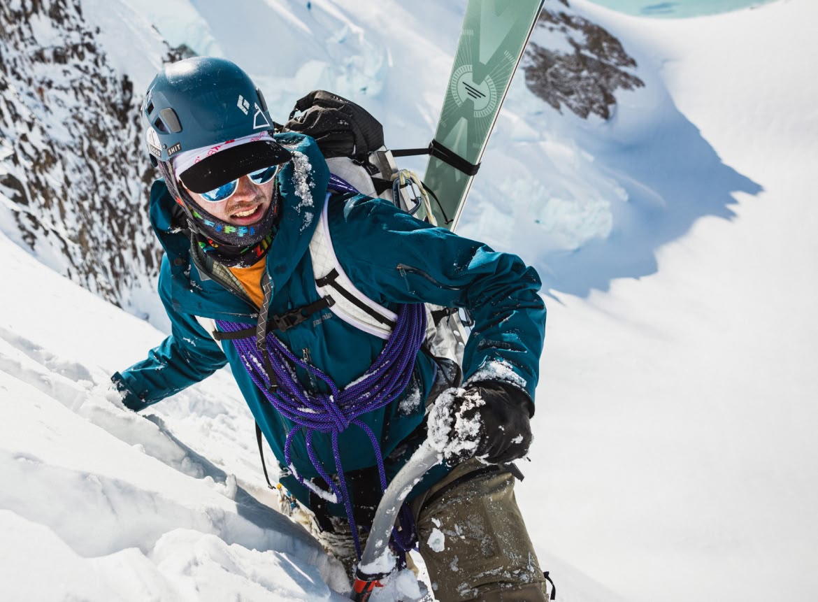 A person wearing a backpack with skis strapped to it climbs a steep, snowy mountain.