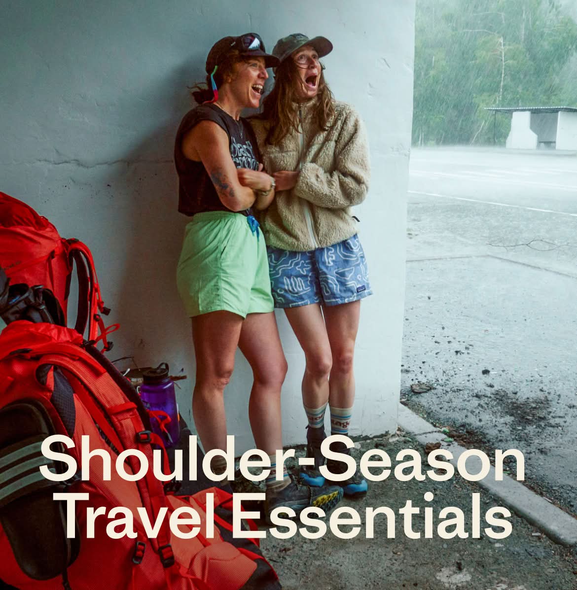 Shoulder-Season Travel Essentials. Two people stand in a bus depot to take shelter from a rain storm. They are laughing and appear amazed at the intensity of the downpour. Their backpacks are stacked in the foreground.