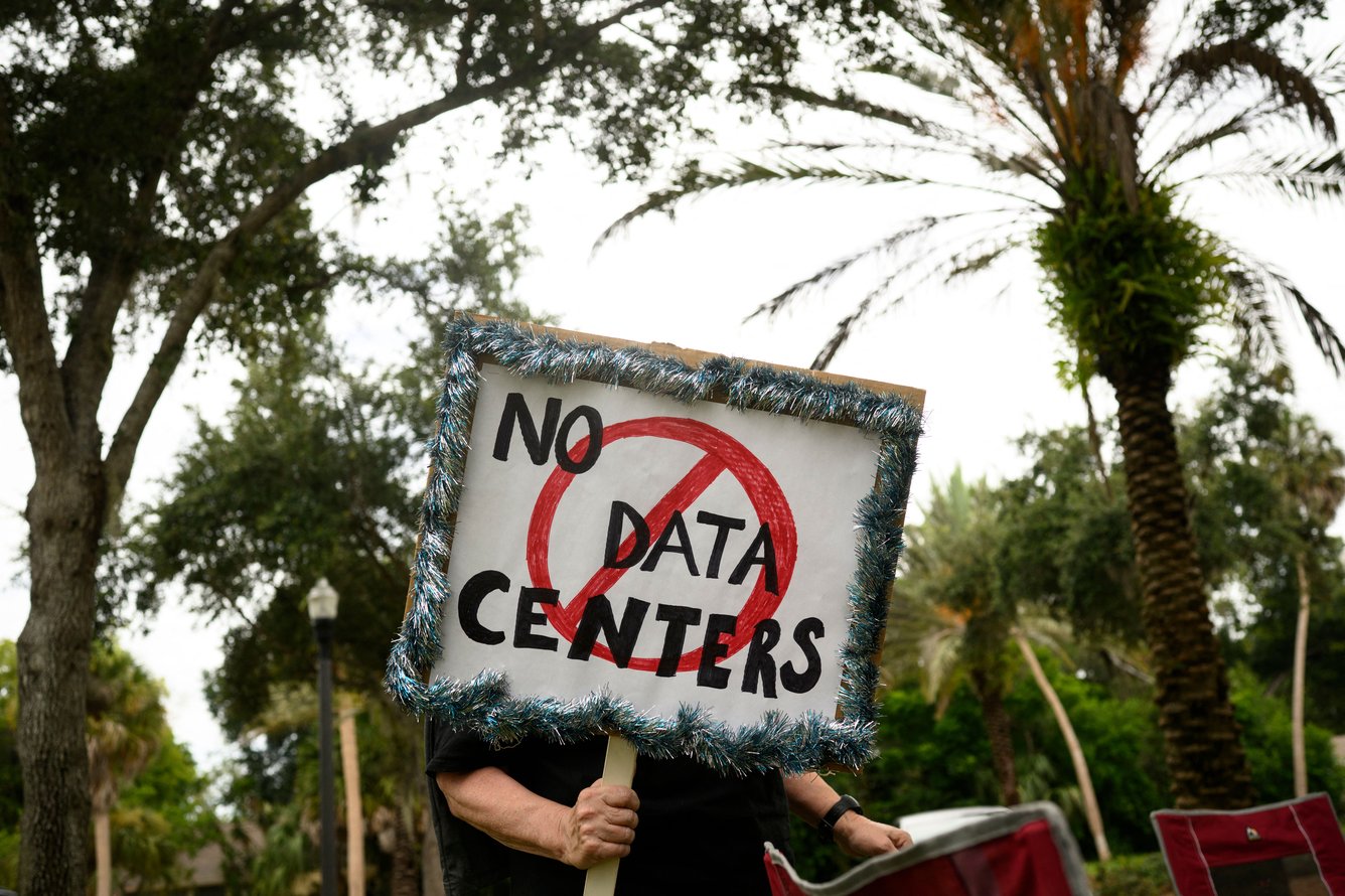 Person holding sign covering their face that says No Data Centers with a red circle ban symbol, standing outside with palm trees in the background.