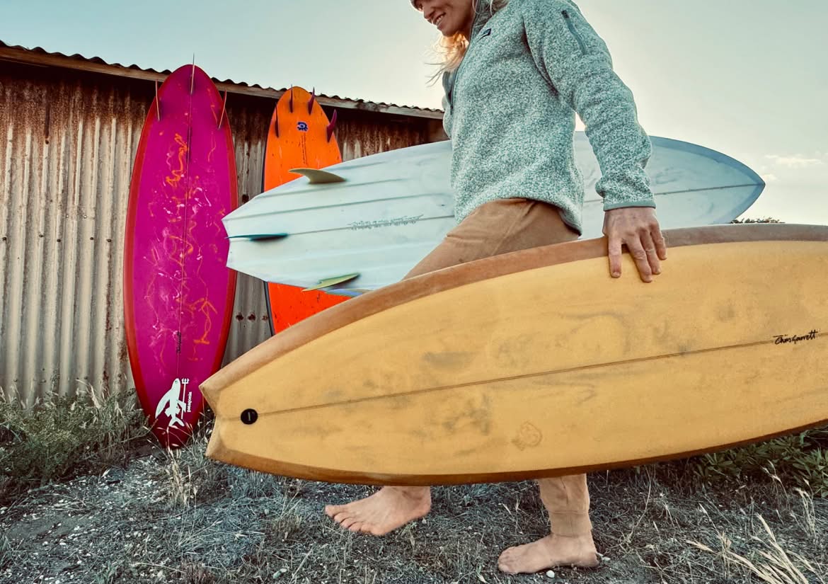 A person carries a surfboard in each arm while walking barefoot past two more surfboards leaning against a sheet metal shed.