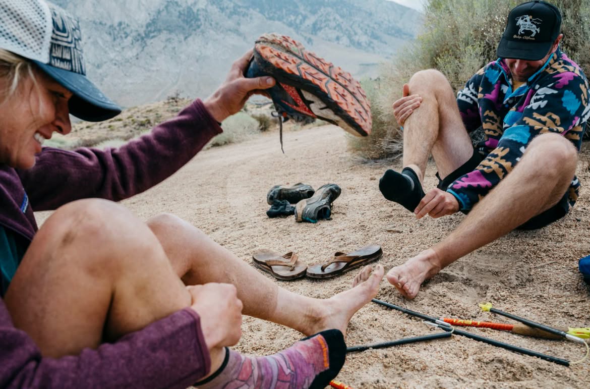 Two people sit on a trail in the mountains and change their shoes and socks. There are a pair of sandals and trekking poles in the dirt between them.