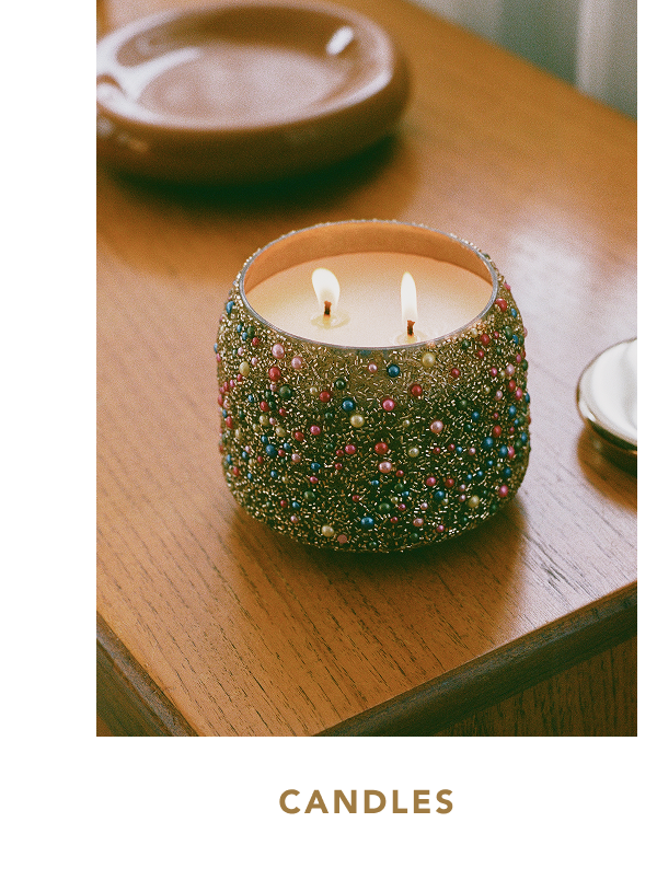 Lit two-wick candle in a cylindrical glass jar fully encrusted with colorful beads and faux pearls, set on a wooden table near a shallow ceramic dish, with text below reading "CANDLES".
