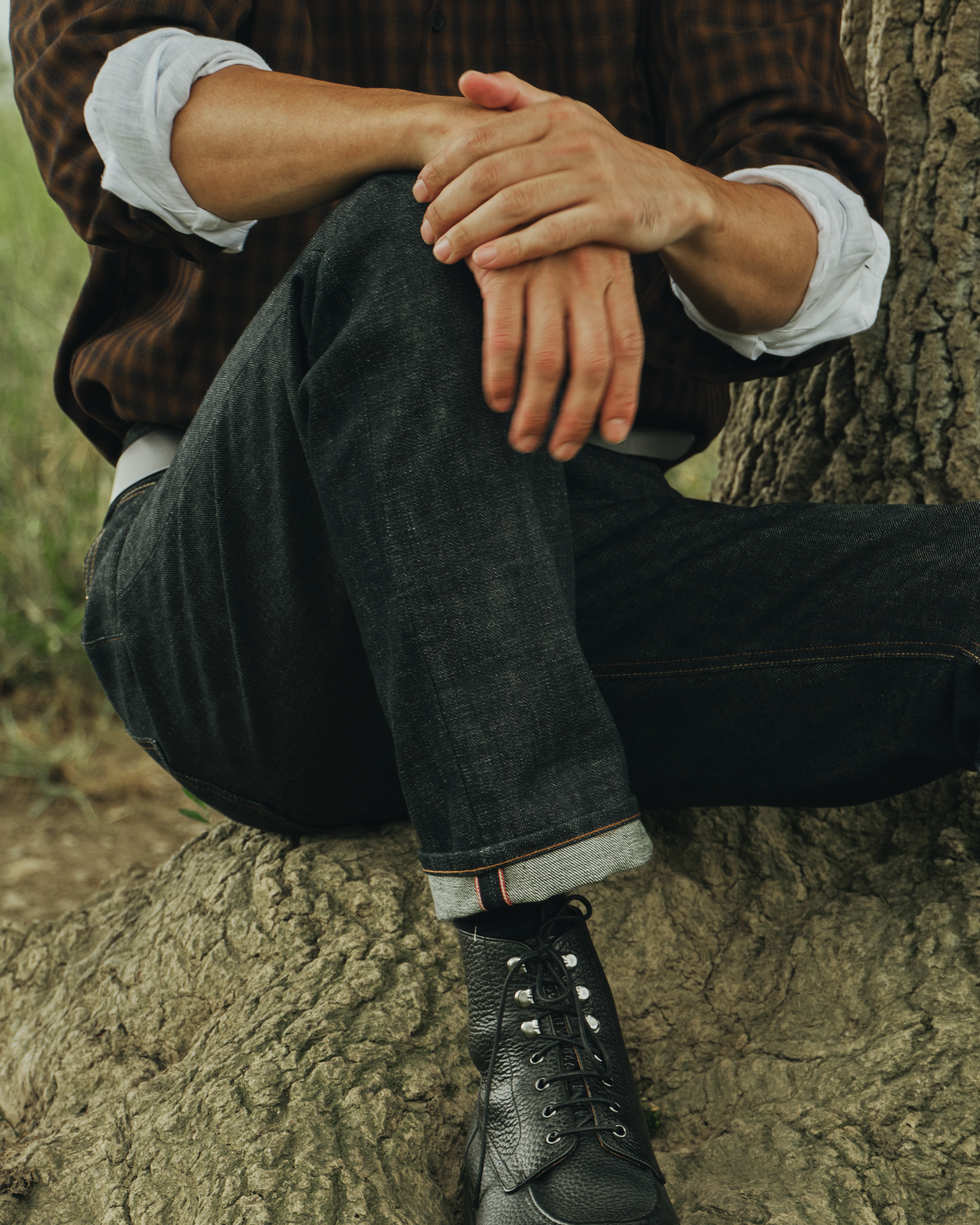 Photo of man sitting at base of tree wearing selvedge jeans, showing small turn-up, black boots and brown shirt.