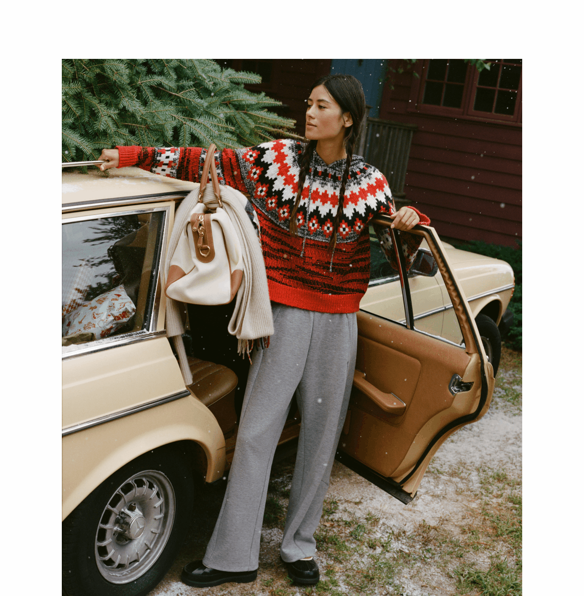 Model standing next to a vintage cream-colored car in a snowy setting, wearing a red, white, and black patterned Fair Isle hoodie sweater and grey wide-leg pants while leaning against the open door holding a travel tote bag with a folded scarf over it.