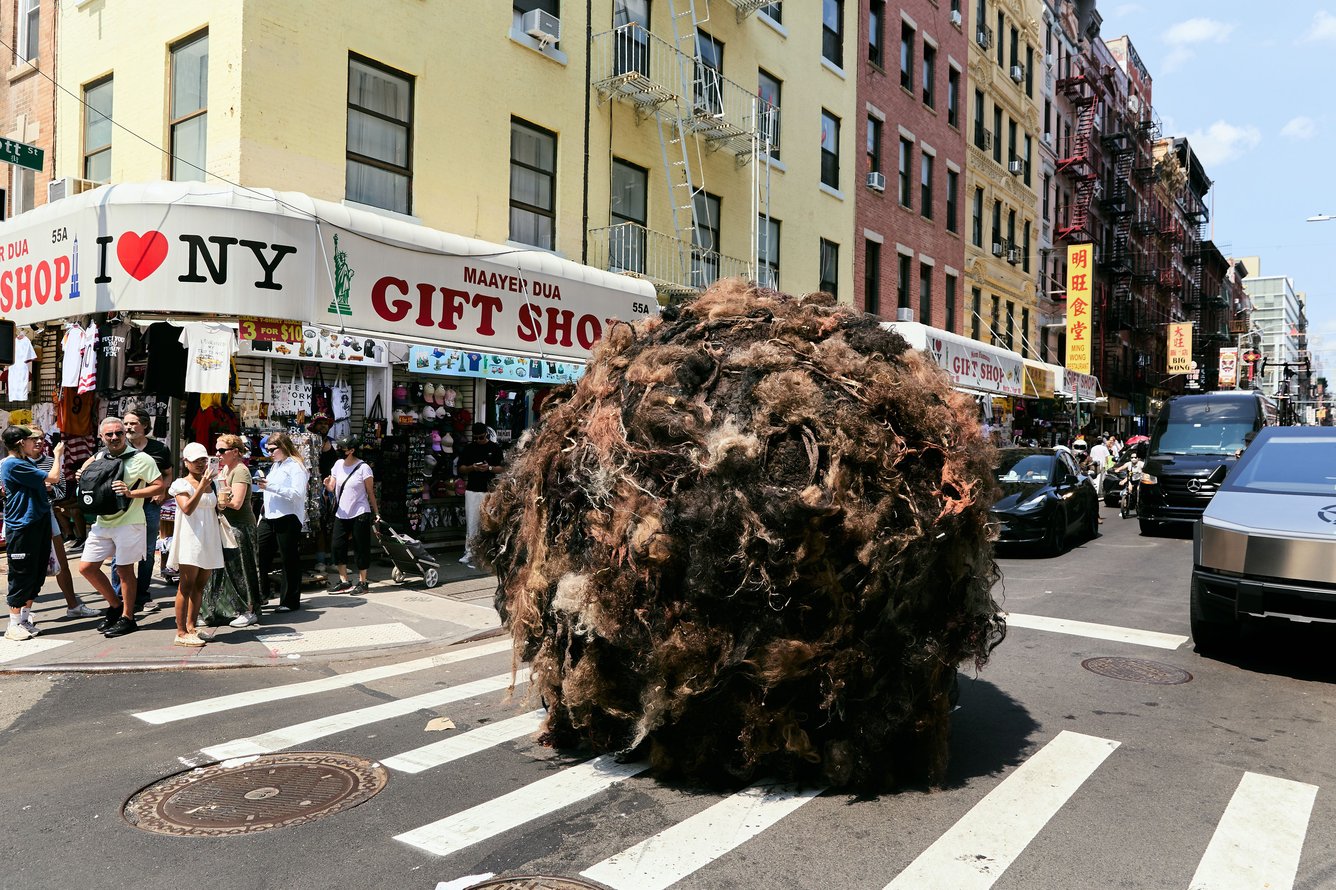 A massive brown ball of human hair, about 10 feet in diameter, in the middle of a crosswalk in New York, with the awning of an NYC gift shop and pedestrians walking visible in the background. 