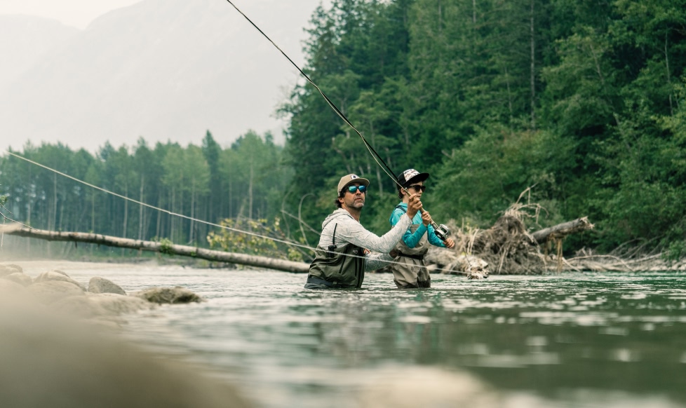Two people wearing waders stand in a river and make a cast with a fly rod. The older person is teaching the younger person how to perform a two-handed cast.