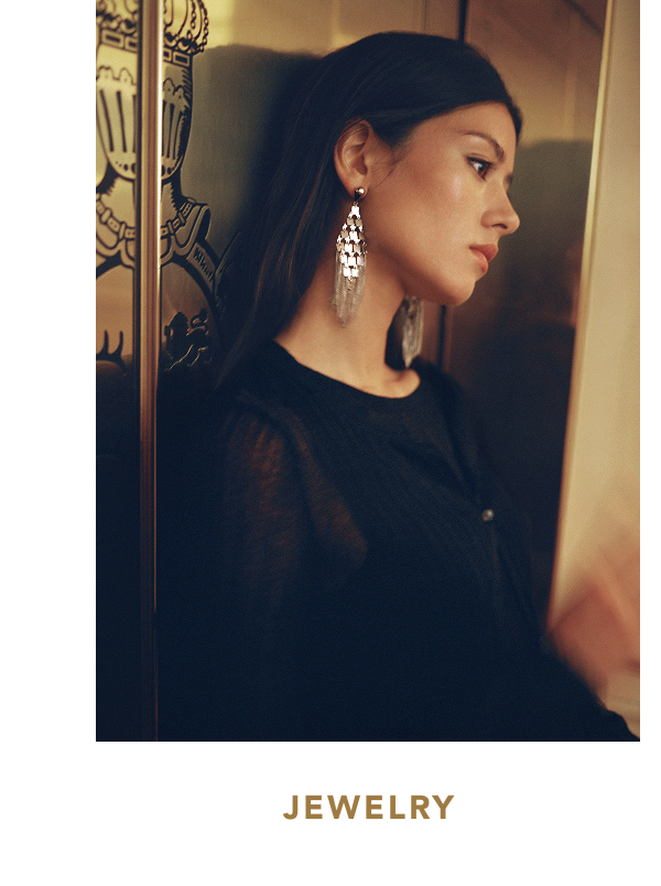 Portrait of a woman leaning against an ornate brass elevator door, wearing a black top and long metallic silver tiered tassel earrings, with text below reading "JEWELRY".