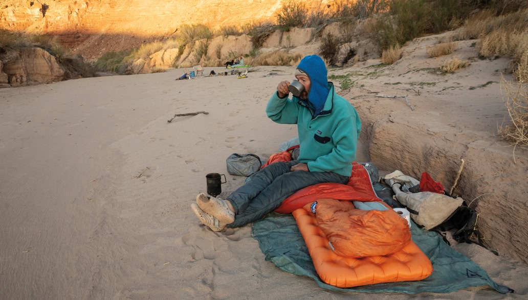 A person sits atop a sleeping bag and inflatable mattress in a sandy river bank and sips a beverage from a camp cup. Two people, a dog and campfire in the background.
