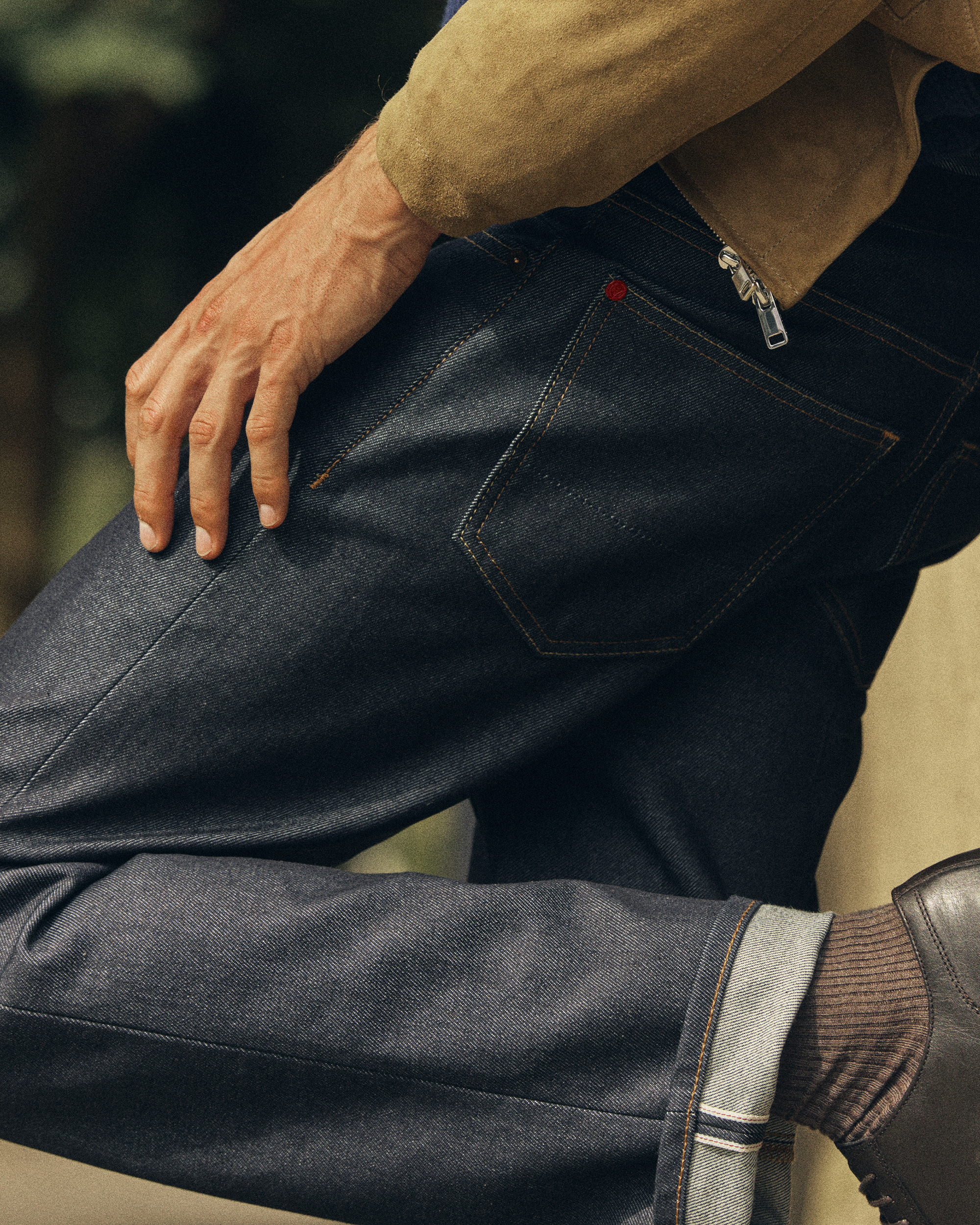 Waist down photo of man leaning against wall wearing Super Zero selvedge Hiut jeans, brown socks, leather shoes, and light brown suede jacket.