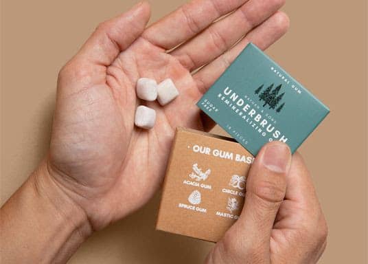 Close-up overhead view of two hands holding three small white gum pieces and overlapping teal and brown rectangular packages against a plain tan backg...