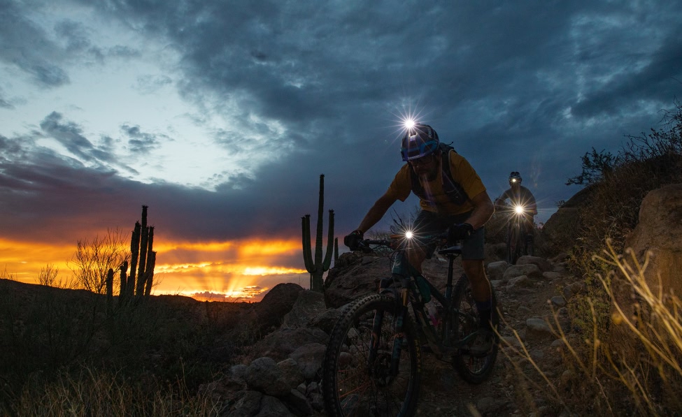 Two mountain bikers wearing headlamps ride down a rocky trail at dusk. The setting sun silhouettes tall cactus and desert hills.