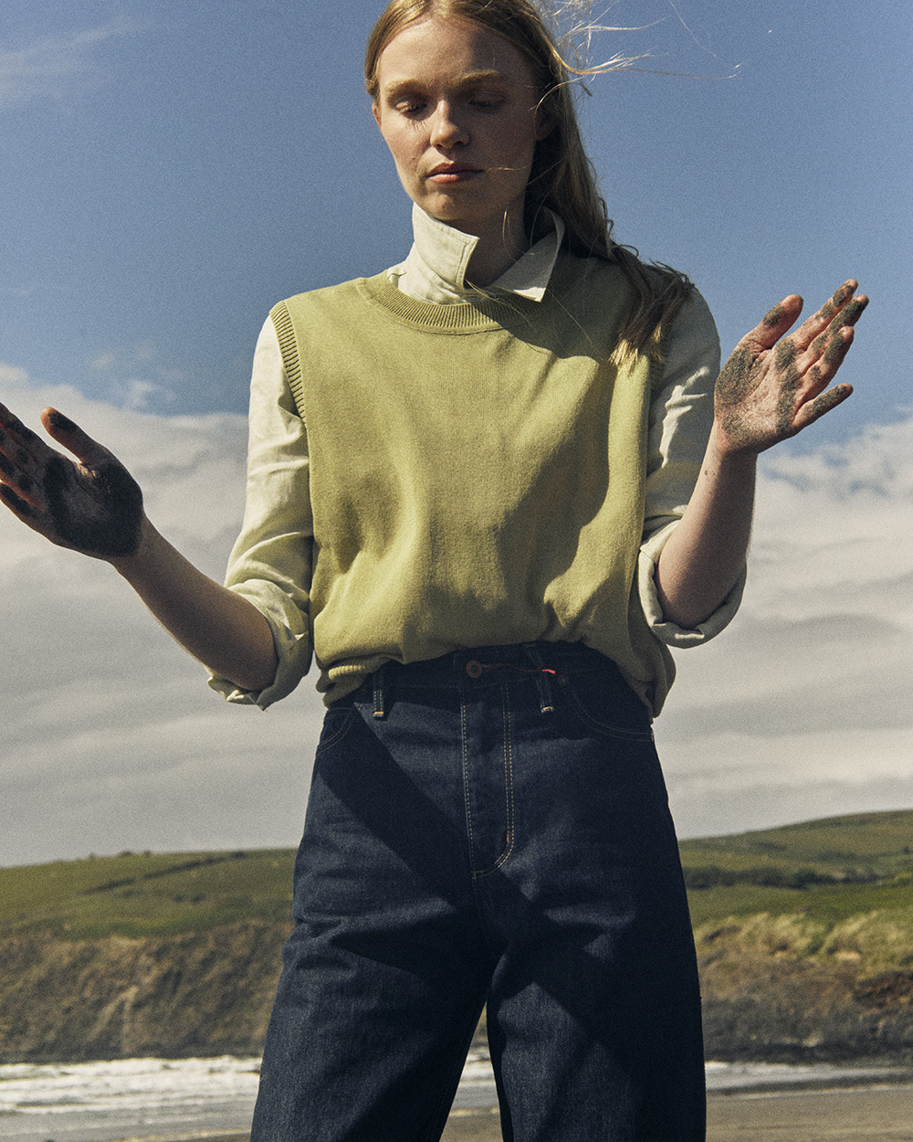 Image of woman on beach with sand covered hands. She wears Rinse Wash denim jeans, green shirt, and green sleeveless jumper.