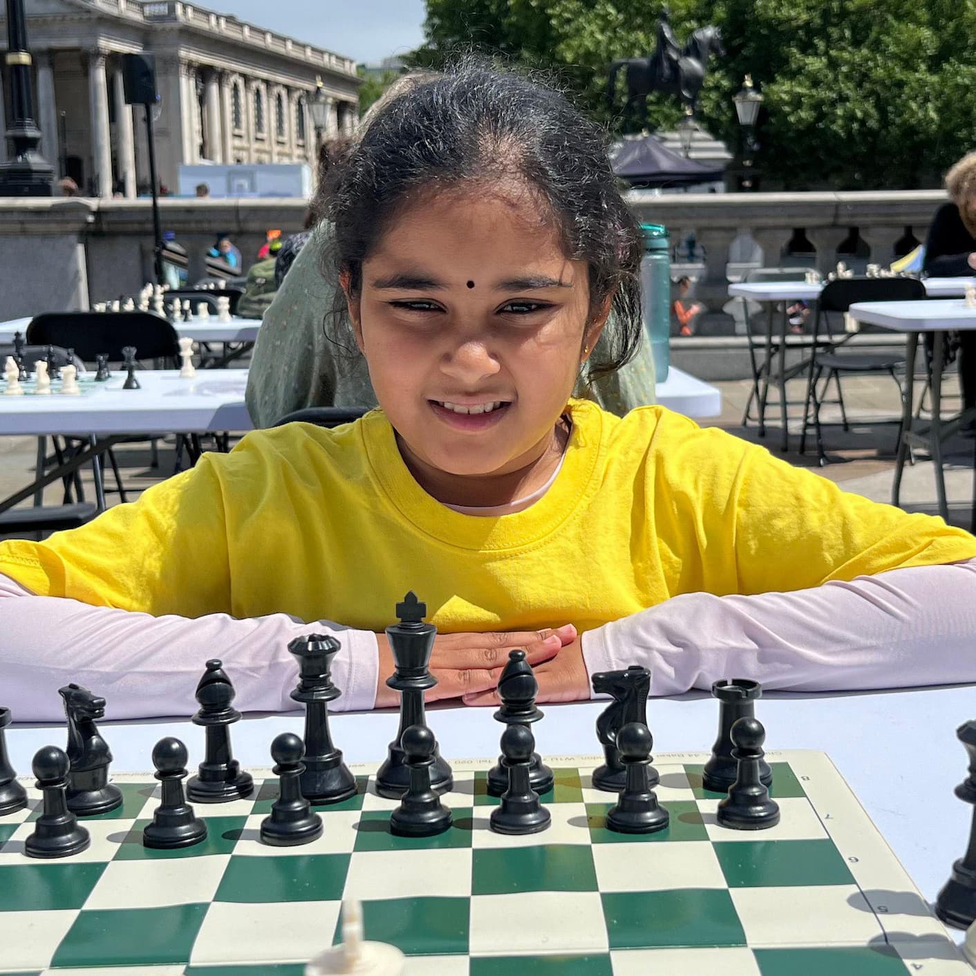 Young chess player in a yellow shirt seated behind a chessboard at an outdoor event.