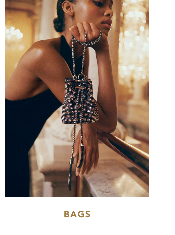 Model wearing a dark halter dress resting her arm on a brass railing, holding up a small black rhinestone mini bucket bag with a chain strap and tassels, with text below reading "BAGS".