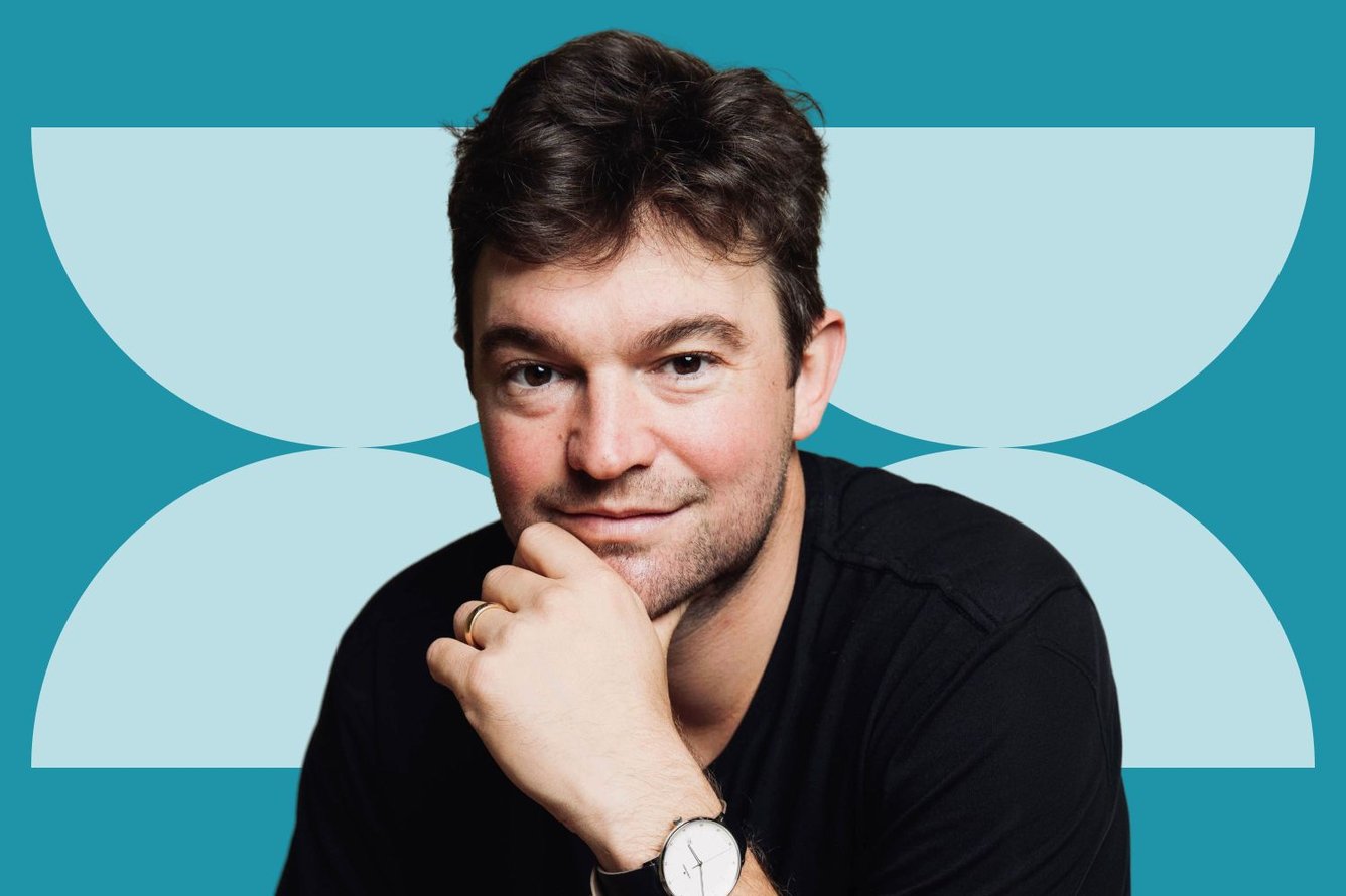 Headshot of Bryant Kohler, a light-skinned man with short wavy dark brown hair and stubble wearing a black shirt, holding his chin in his hand thoughtfully while smiling at the camera.