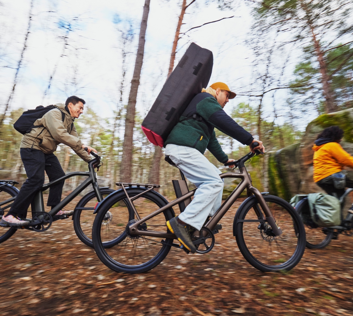 Riders on Cowboy Cross e-bikes on a forest trail, one carrying a large bouldering crash pad on his back.